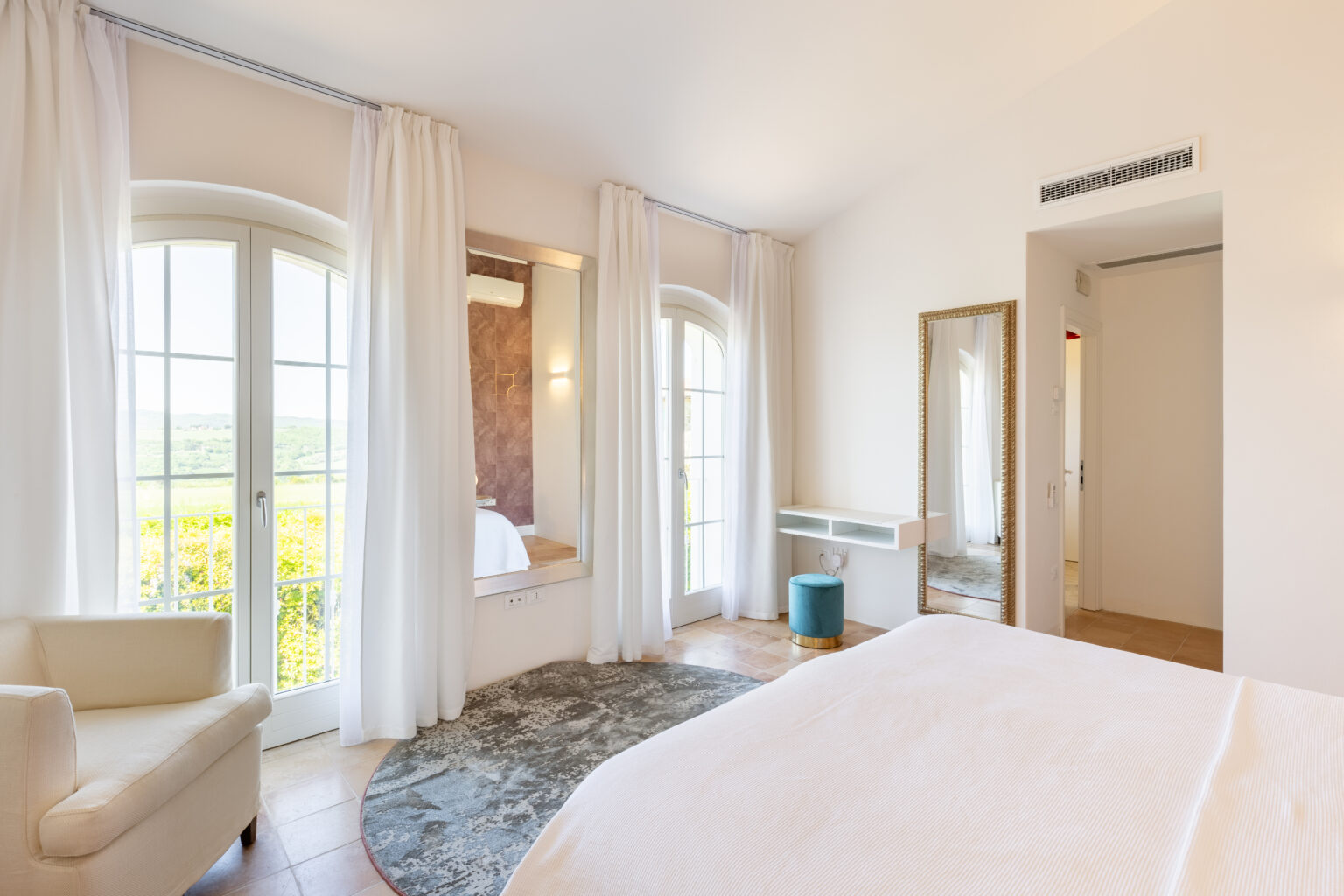 Bright and airy bedroom featuring large windows with sheer curtains, a comfortable bed, a cozy armchair, and a stylish mirror. The room is decorated in neutral tones with a soft area rug, creating a serene atmosphere and offering scenic views of the outdoors. Ideal for relaxation and comfort. | Only on SopranoVillas