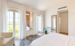 Bright and airy bedroom featuring large windows with sheer curtains, a comfortable bed, a cozy armchair, and a stylish mirror. The room is decorated in neutral tones with a soft area rug, creating a serene atmosphere and offering scenic views of the outdoors. Ideal for relaxation and comfort.