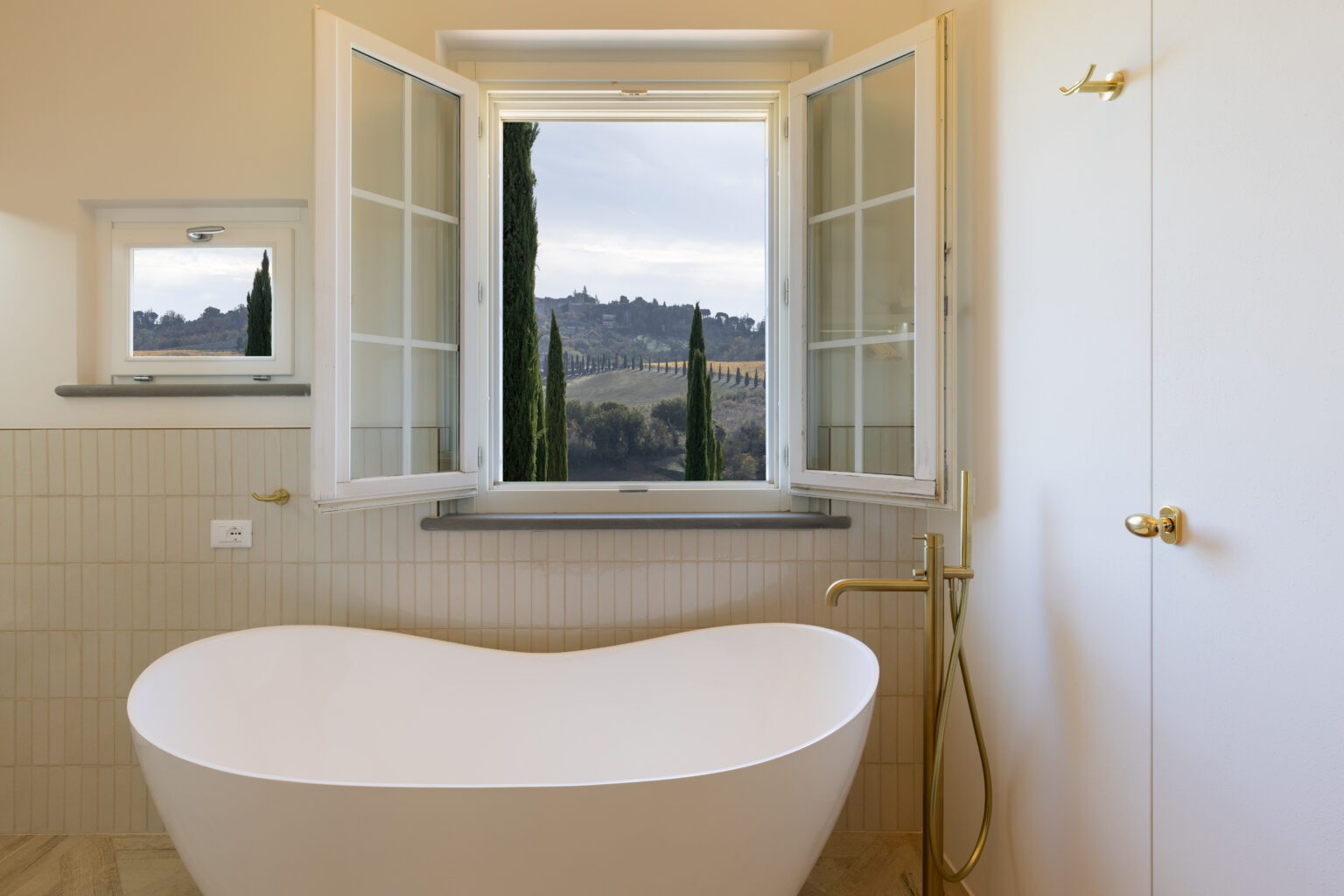 Modern bathroom featuring a freestanding white bathtub, gold fixtures, and large windows offering a scenic view of rolling hills and cypress trees. The neutral tile design enhances the serene ambiance. | Only on SopranoVillas