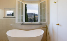 Modern bathroom featuring a freestanding white bathtub, gold fixtures, and large windows offering a scenic view of rolling hills and cypress trees. The neutral tile design enhances the serene ambiance.