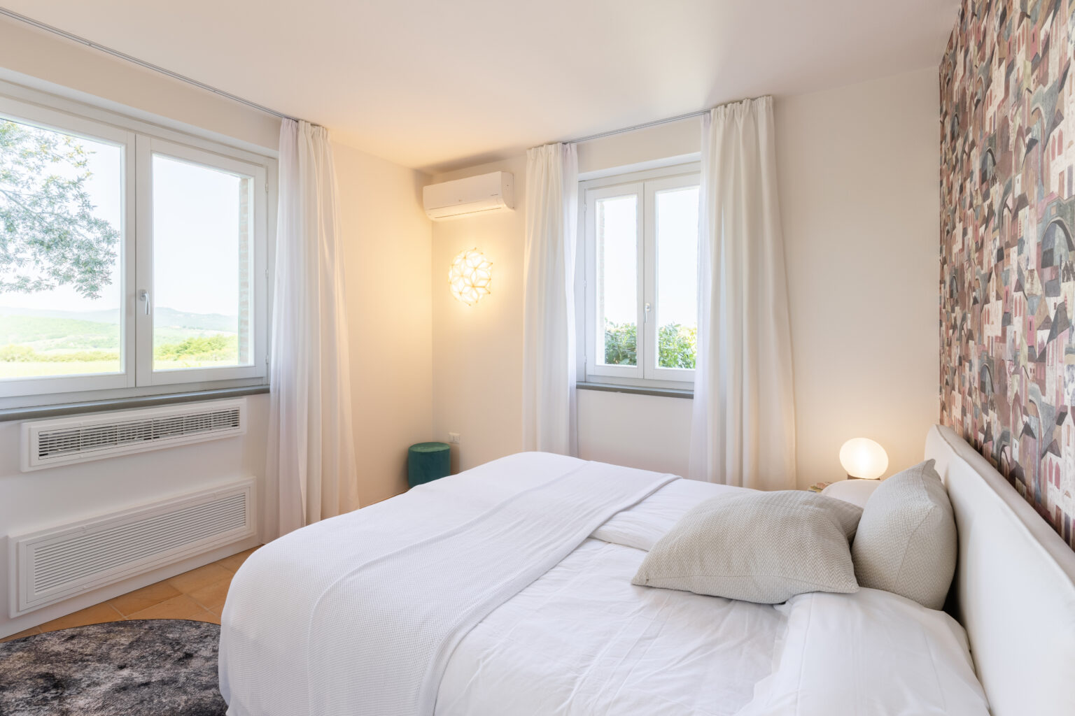 Bright and airy bedroom featuring a comfortable bed with decorative pillows, large windows with sheer curtains, and a stylish accent wall. The room is illuminated by a modern light fixture and includes a cozy rug and a small side table, offering a serene and inviting atmosphere. | Only on SopranoVillas