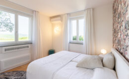 Bright and airy bedroom featuring a comfortable bed with decorative pillows, large windows with sheer curtains, and a stylish accent wall. The room is illuminated by a modern light fixture and includes a cozy rug and a small side table, offering a serene and inviting atmosphere.