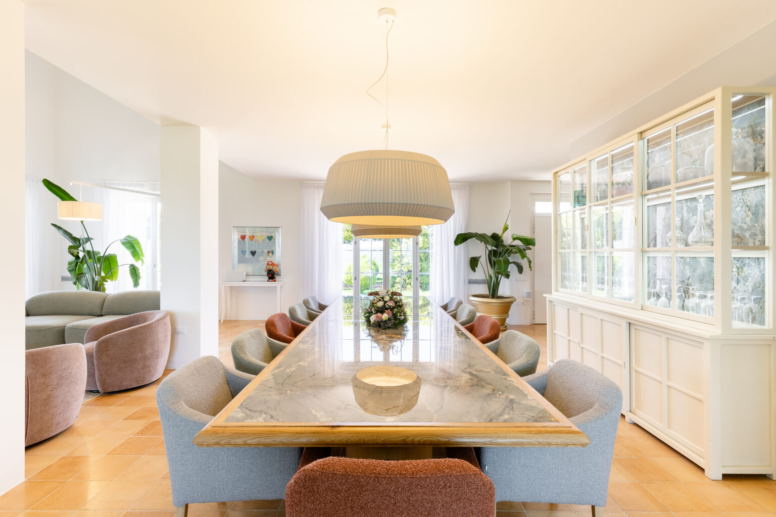 Modern dining room featuring a long marble table surrounded by stylish upholstered chairs in various colors, complemented by a large pendant light and a decorative cabinet. Lush greenery and a bright, airy atmosphere enhance the inviting space. | Only on SopranoVillas