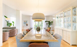 Modern dining room featuring a long marble table surrounded by stylish upholstered chairs in various colors, complemented by a large pendant light and a decorative cabinet. Lush greenery and a bright, airy atmosphere enhance the inviting space.
