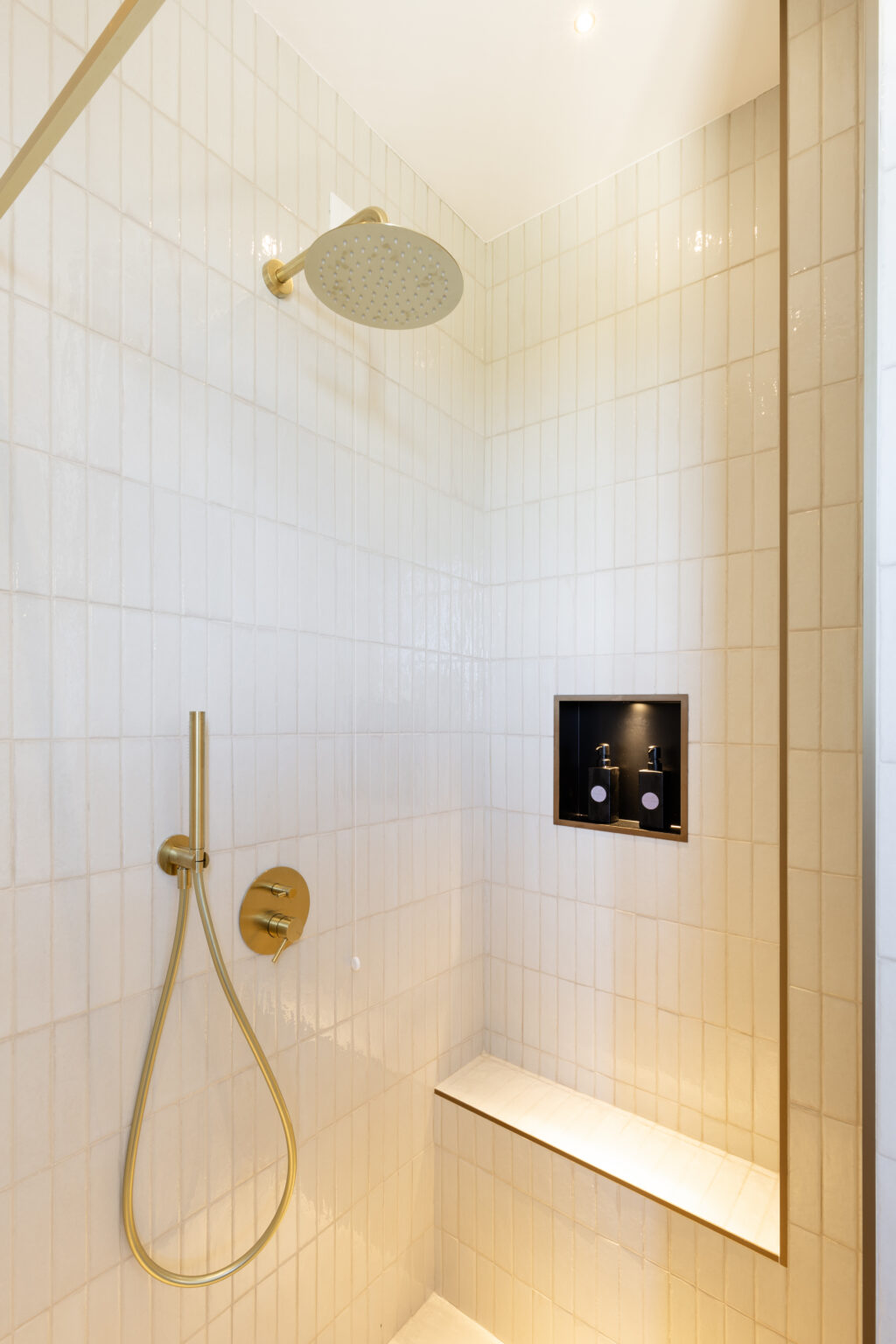 Modern shower design featuring a gold rain showerhead, sleek handheld faucet, and built-in niche for toiletries against a backdrop of white tiled walls. The spacious layout includes a built-in bench for added comfort. | Only on SopranoVillas
