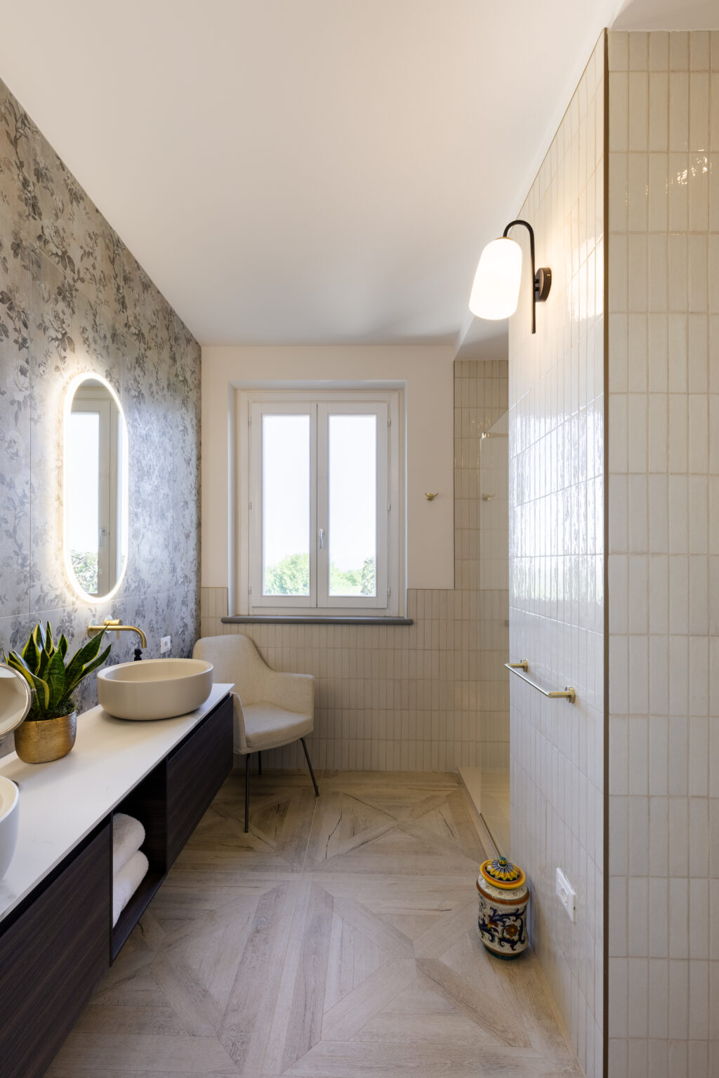 Modern bathroom interior featuring a stylish design with a floral wallpaper accent, round backlit mirror, elegant vessel sinks, and a cozy chair. Natural light streams in through a window, highlighting the textured wall tiles and wooden flooring. | Only on SopranoVillas