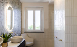 Modern bathroom interior featuring a stylish design with a floral wallpaper accent, round backlit mirror, elegant vessel sinks, and a cozy chair. Natural light streams in through a window, highlighting the textured wall tiles and wooden flooring.