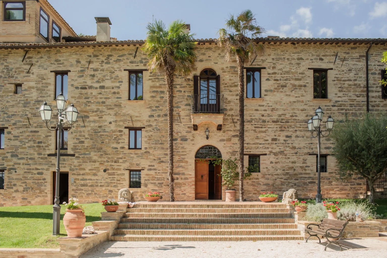 Historic stone building with palm trees, a wooden door, and decorative flower pots, set in a garden with steps leading to the entrance. | Only on SopranoVillas