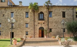 Historic stone building with palm trees, a wooden door, and decorative flower pots, set in a garden with steps leading to the entrance.