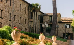 Historic stone building with landscaped gardens and decorative planters filled with flowers, set against a clear blue sky. The scene features a gravel pathway leading to the entrance, surrounded by lush greenery and tall trees, creating a serene and inviting atmosphere.