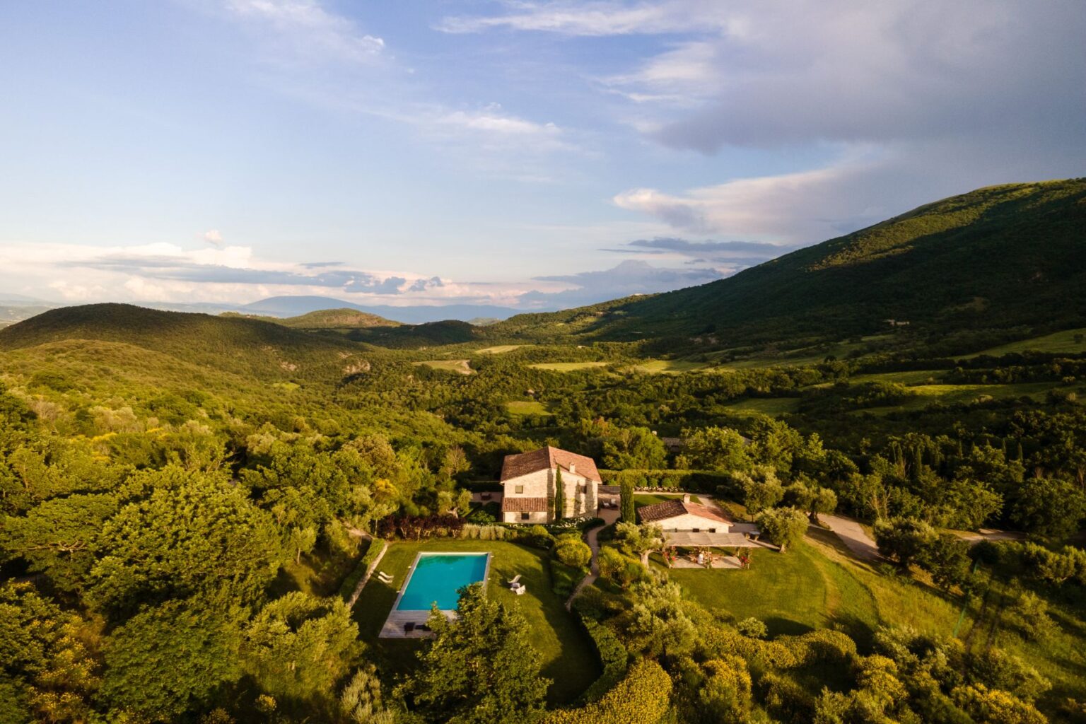 Aerial view of a picturesque countryside villa surrounded by lush green hills, featuring a swimming pool and landscaped gardens, ideal for relaxation and retreat. | Only on SopranoVillas