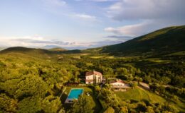 Aerial view of a picturesque countryside villa surrounded by lush green hills, featuring a swimming pool and landscaped gardens, ideal for relaxation and retreat.
