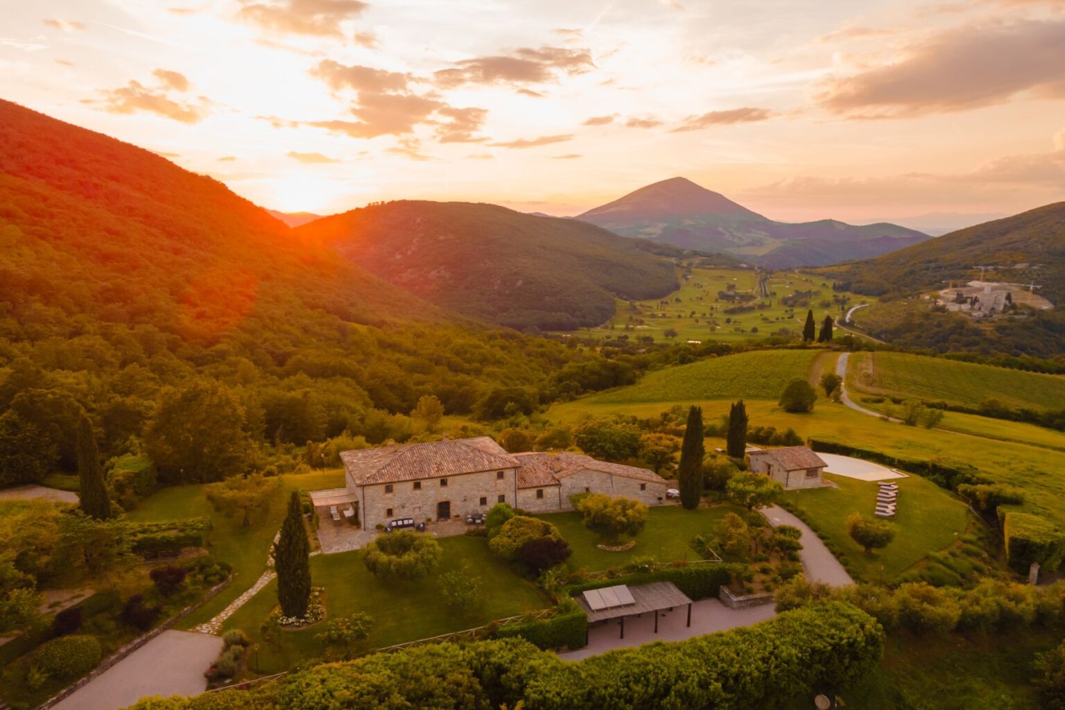 Scenic aerial view of a rustic stone villa surrounded by lush greenery and rolling hills at sunset, with mountains in the background and a winding road leading through the countryside. | Only on SopranoVillas