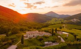 Scenic aerial view of a rustic stone villa surrounded by lush greenery and rolling hills at sunset, with mountains in the background and a winding road leading through the countryside.