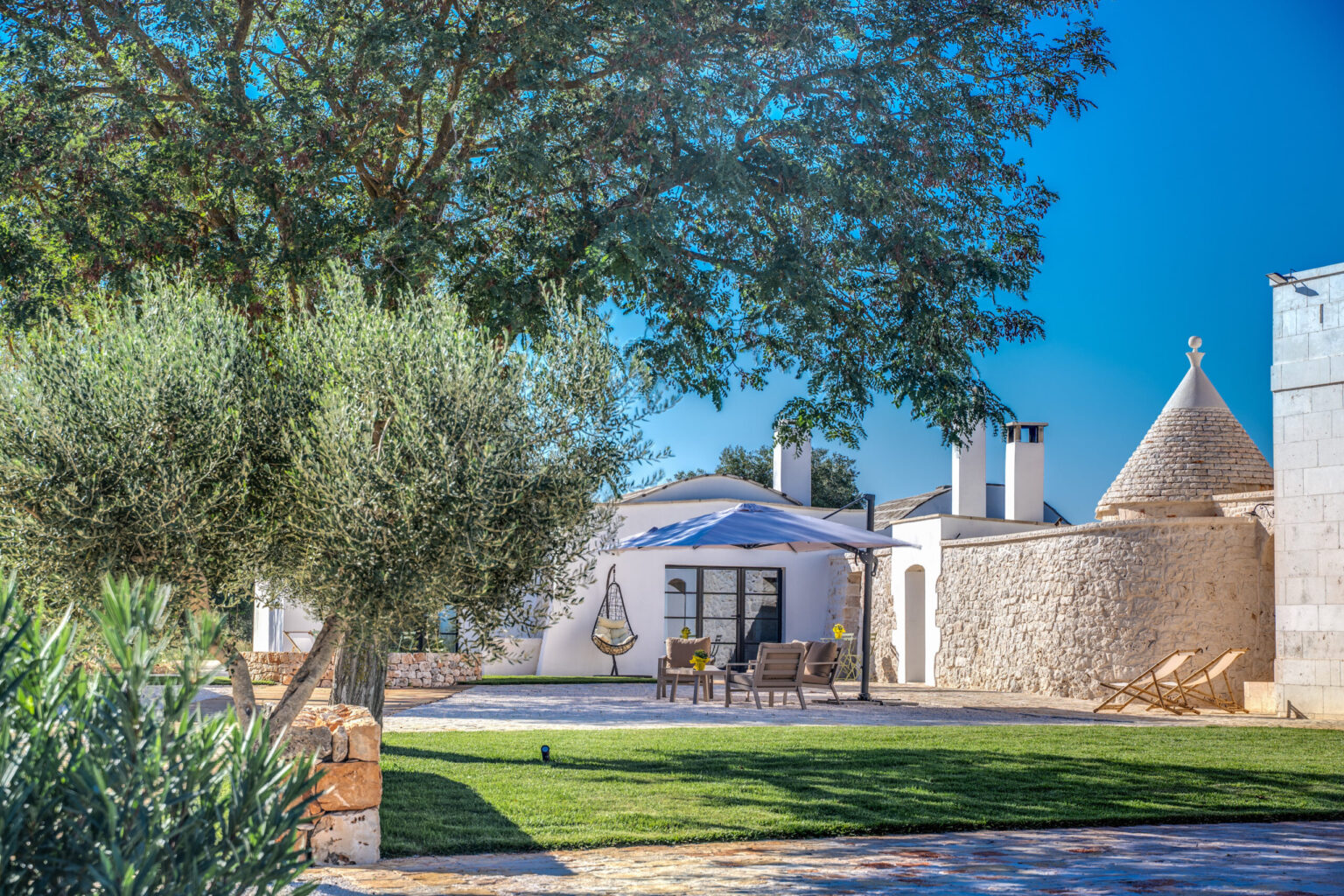 Tranquil outdoor setting featuring a white stone villa with a conical roof, surrounded by olive trees and lush green grass. A shaded seating area with modern furniture provides a relaxing atmosphere under a clear blue sky. Ideal for vacation rentals or serene getaways. | Only on SopranoVillas