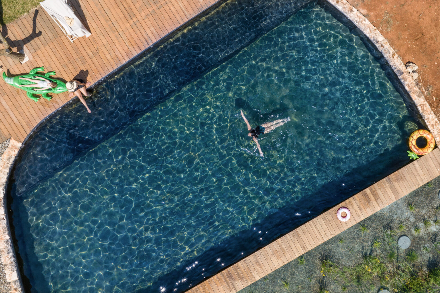 Aerial view of a serene swimming pool with a person floating on their back, surrounded by clear blue water. A wooden deck lines the pool's edge, featuring a green inflatable alligator and a colorful pool float nearby. Sunlight reflects off the water, creating a tranquil atmosphere ideal for relaxation and leisure. | Only on SopranoVillas