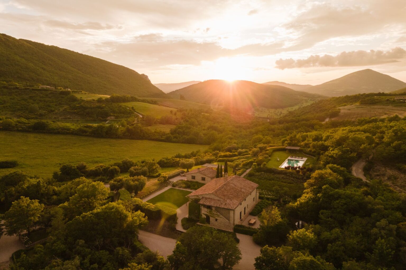 Aerial view of a rustic villa surrounded by lush green hills and fields at sunset, showcasing a swimming pool and landscaped gardens in a serene countryside setting. | Only on SopranoVillas