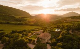 Aerial view of a rustic villa surrounded by lush green hills and fields at sunset, showcasing a swimming pool and landscaped gardens in a serene countryside setting.