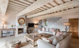 Cozy living room featuring exposed wooden beams, a stone fireplace, and stylish furnishings. The space includes a flat-screen TV, decorative accents, and a natural fiber rug, creating a warm and inviting atmosphere.