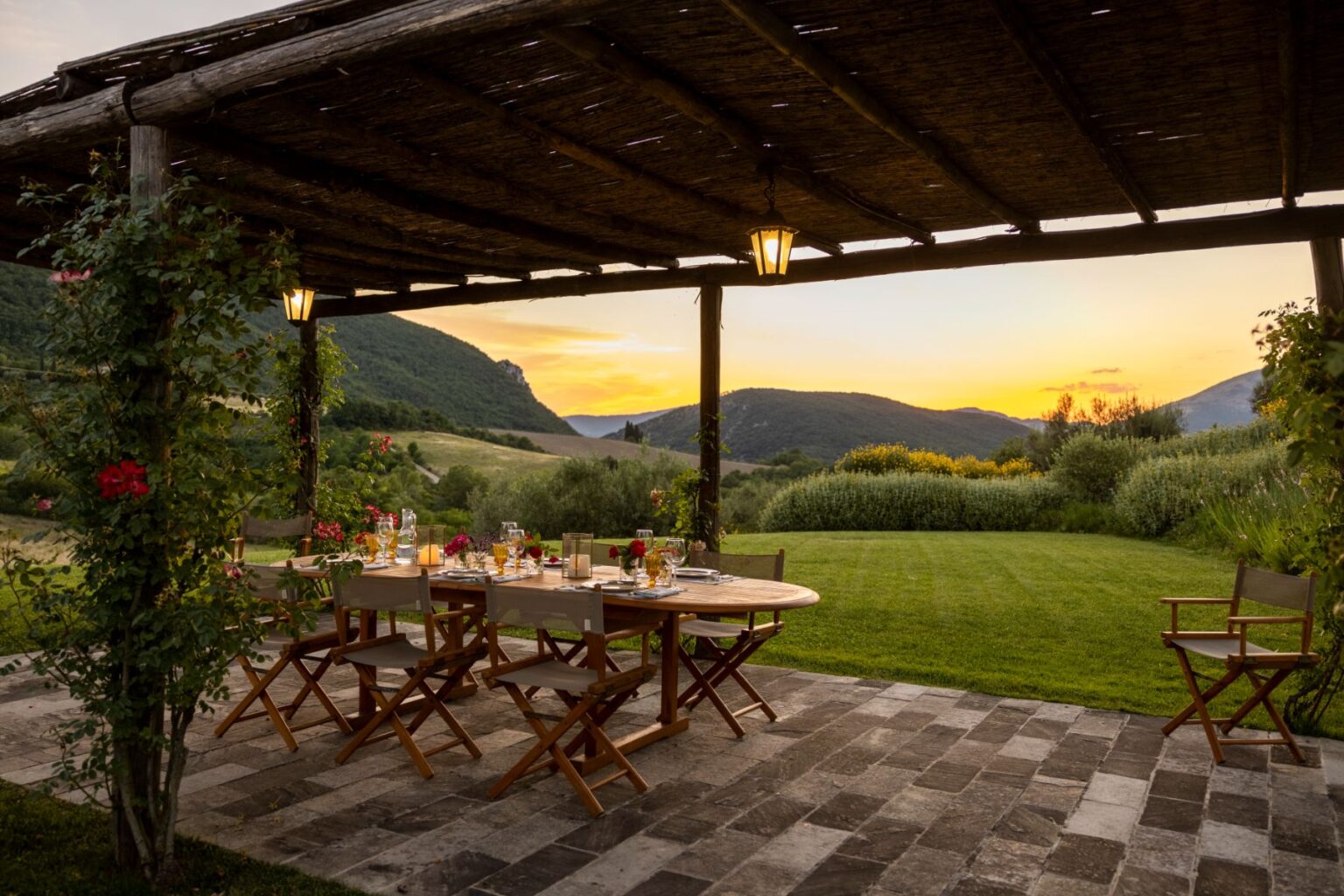 Outdoor dining area under a rustic pergola, featuring a wooden table set for a meal with elegant glassware and floral decorations. The backdrop includes rolling hills and a vibrant sunset, creating a picturesque and inviting atmosphere for gatherings. | Only on SopranoVillas