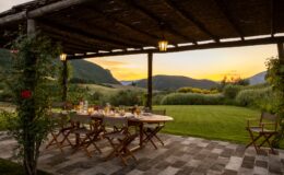 Outdoor dining area under a rustic pergola, featuring a wooden table set for a meal with elegant glassware and floral decorations. The backdrop includes rolling hills and a vibrant sunset, creating a picturesque and inviting atmosphere for gatherings.