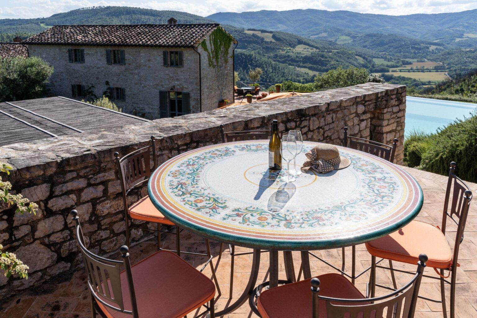 Scenic outdoor dining area featuring a decorative tiled table set with a bottle of wine and glasses, surrounded by stone walls and overlooking lush green hills and a swimming pool. Perfect for enjoying meals in a tranquil countryside setting. | Only on SopranoVillas