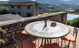 Scenic outdoor dining area featuring a decorative tiled table set with a bottle of wine and glasses, surrounded by stone walls and overlooking lush green hills and a swimming pool. Perfect for enjoying meals in a tranquil countryside setting.