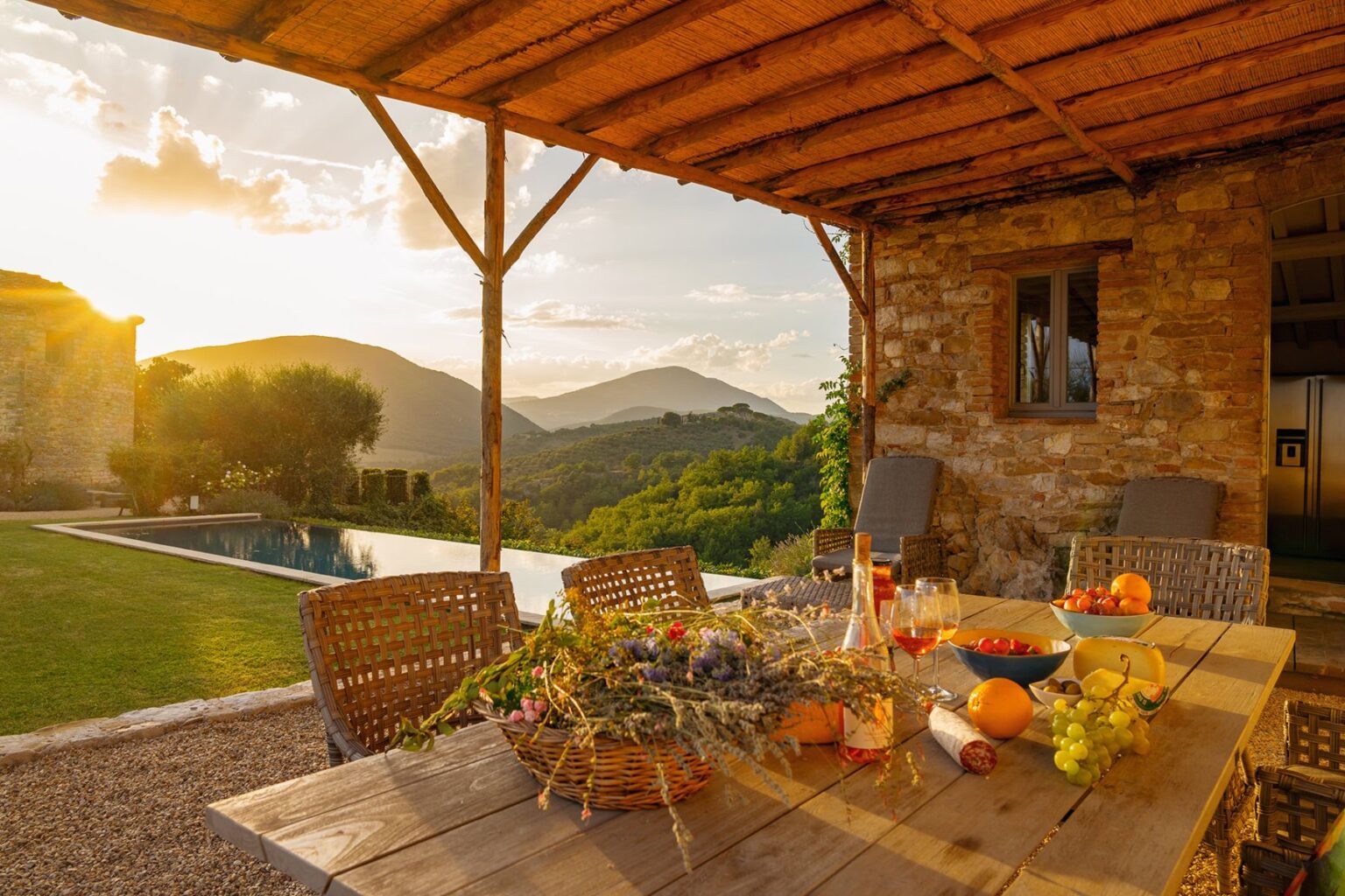 Beautiful outdoor dining area with a wooden table set with fresh fruits and drinks, surrounded by a scenic view of mountains and a swimming pool at sunset. The rustic stone building and lush greenery enhance the serene atmosphere. | Only on SopranoVillas
