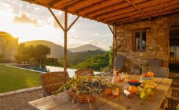 Beautiful outdoor dining area with a wooden table set with fresh fruits and drinks, surrounded by a scenic view of mountains and a swimming pool at sunset. The rustic stone building and lush greenery enhance the serene atmosphere.