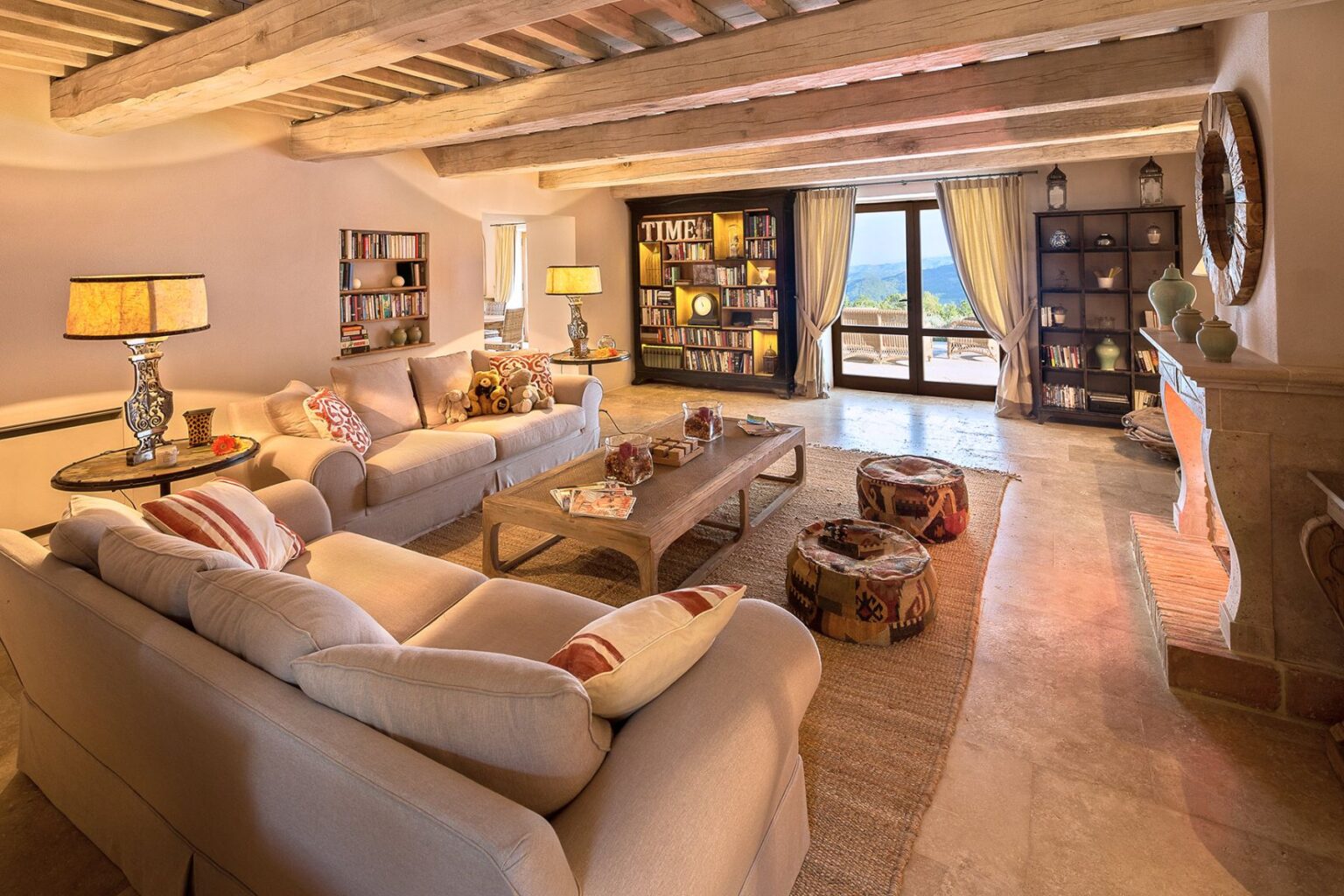 Cozy living room featuring beige sofas with decorative pillows, a wooden coffee table with magazines, and a stylish lamp. A bookshelf filled with books is visible on the wall, while large windows offer scenic views. The room has a warm, inviting atmosphere with rustic wooden beams and stone flooring. | Only on SopranoVillas