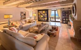 Cozy living room featuring beige sofas with decorative pillows, a wooden coffee table with magazines, and a stylish lamp. A bookshelf filled with books is visible on the wall, while large windows offer scenic views. The room has a warm, inviting atmosphere with rustic wooden beams and stone flooring.