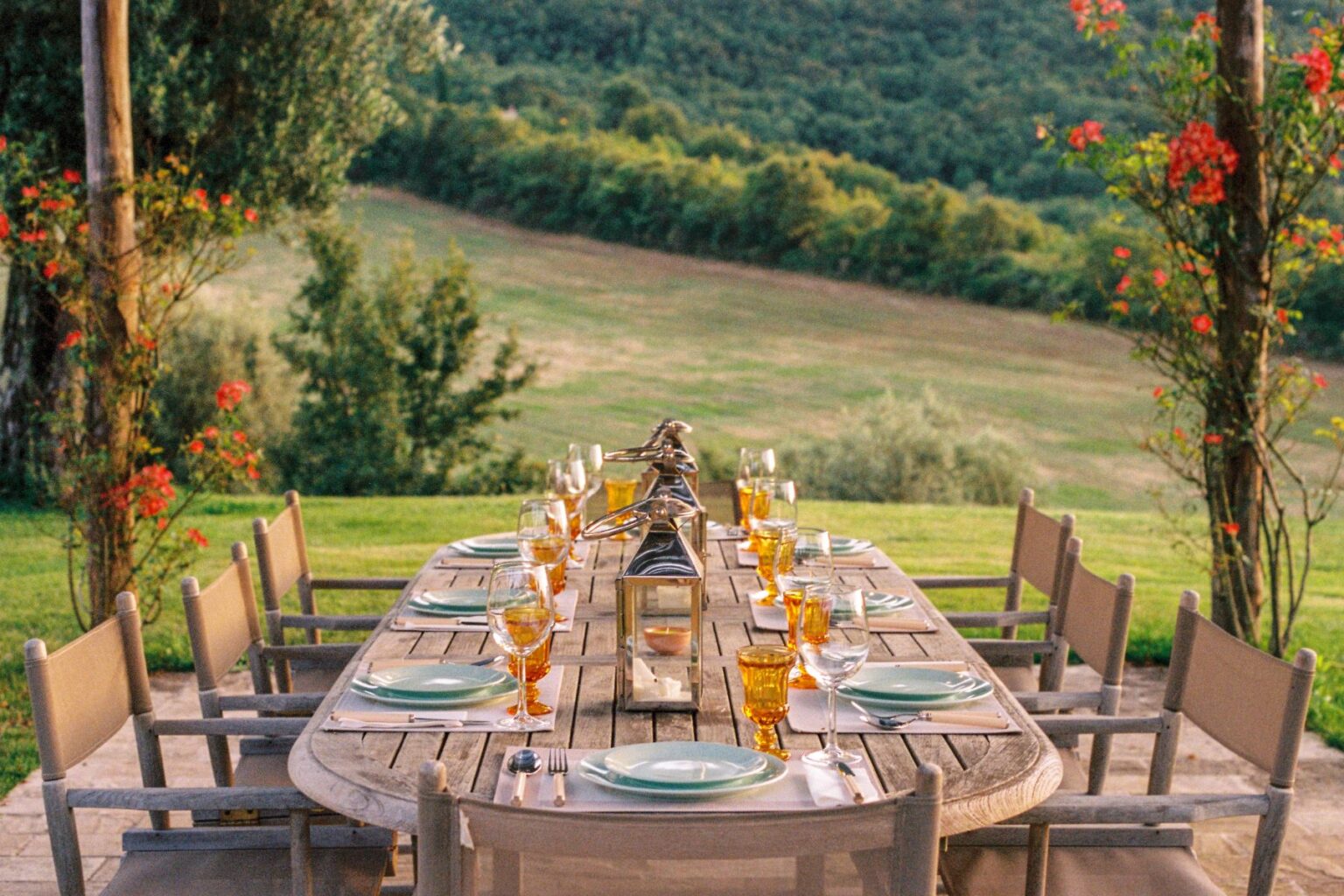 Outdoor dining setup on a wooden table with elegant glassware and plates, surrounded by lush greenery and flowers, creating a picturesque setting for a gathering or celebration. | Only on SopranoVillas