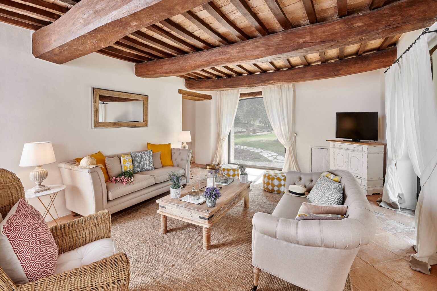 Cozy living room featuring rustic wooden beams, plush sofas with colorful cushions, a wooden coffee table, and a large window offering a view of the outdoors. Decor includes a stylish lamp, potted plants, and a flat-screen TV against a light-colored wall. | Only on SopranoVillas
