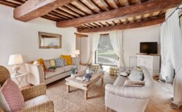 Cozy living room featuring rustic wooden beams, plush sofas with colorful cushions, a wooden coffee table, and a large window offering a view of the outdoors. Decor includes a stylish lamp, potted plants, and a flat-screen TV against a light-colored wall.