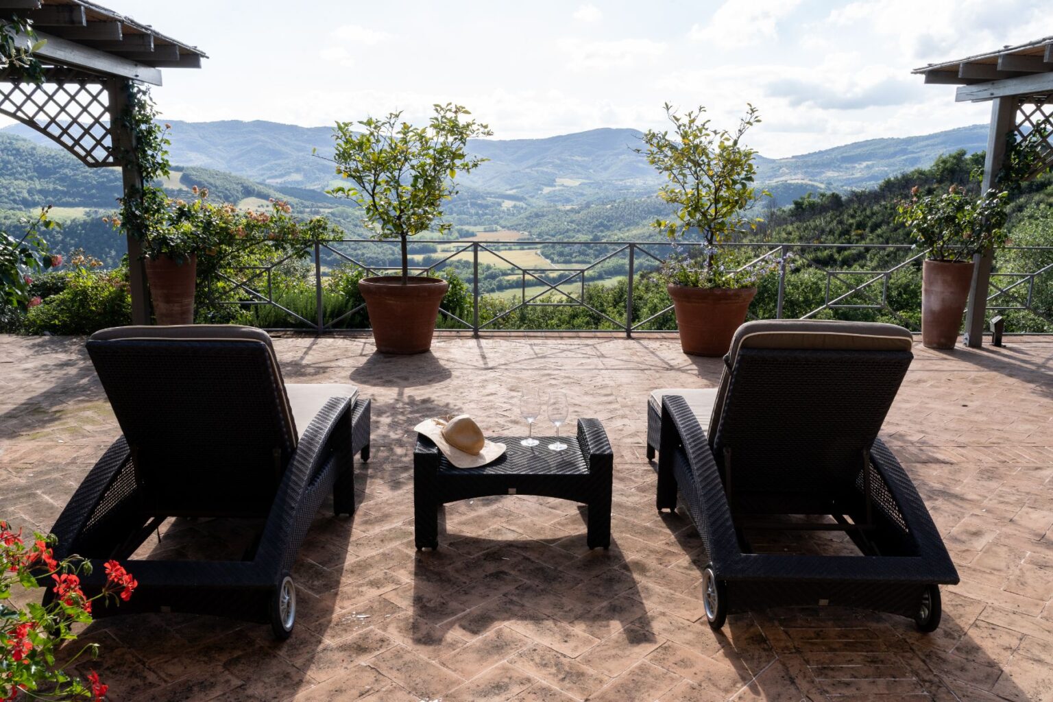Two lounge chairs on a patio overlooking a scenic landscape with rolling hills and greenery. A small table holds a straw hat and two glasses, surrounded by potted plants. Ideal for relaxation and enjoying nature. | Only on SopranoVillas