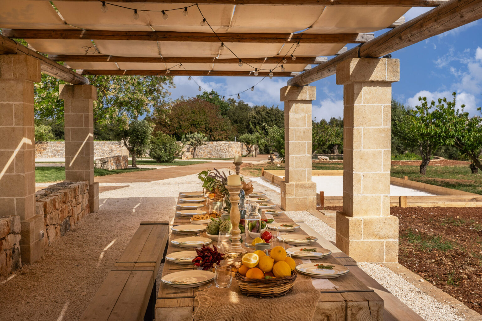 Outdoor dining setup under a pergola with a rustic wooden table, elegantly arranged with plates, glassware, and a centerpiece of fresh fruits and flowers, surrounded by lush greenery and a scenic landscape. | Only on SopranoVillas