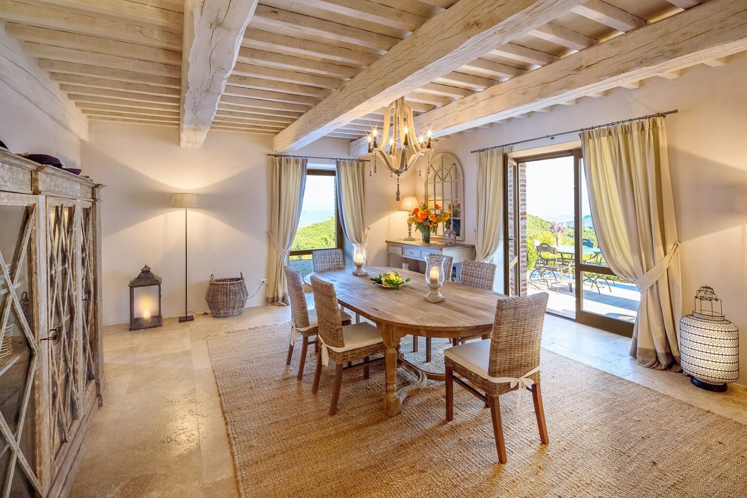 Bright and inviting dining room with rustic wooden beams, a large wooden table surrounded by woven chairs, and elegant decor. Natural light floods the space through large windows, showcasing a scenic view. Features a stylish chandelier, decorative flowers, and a cozy ambiance with soft textures and warm colors. Ideal for family gatherings and entertaining. | Only on SopranoVillas