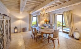 Bright and inviting dining room with rustic wooden beams, a large wooden table surrounded by woven chairs, and elegant decor. Natural light floods the space through large windows, showcasing a scenic view. Features a stylish chandelier, decorative flowers, and a cozy ambiance with soft textures and warm colors. Ideal for family gatherings and entertaining.