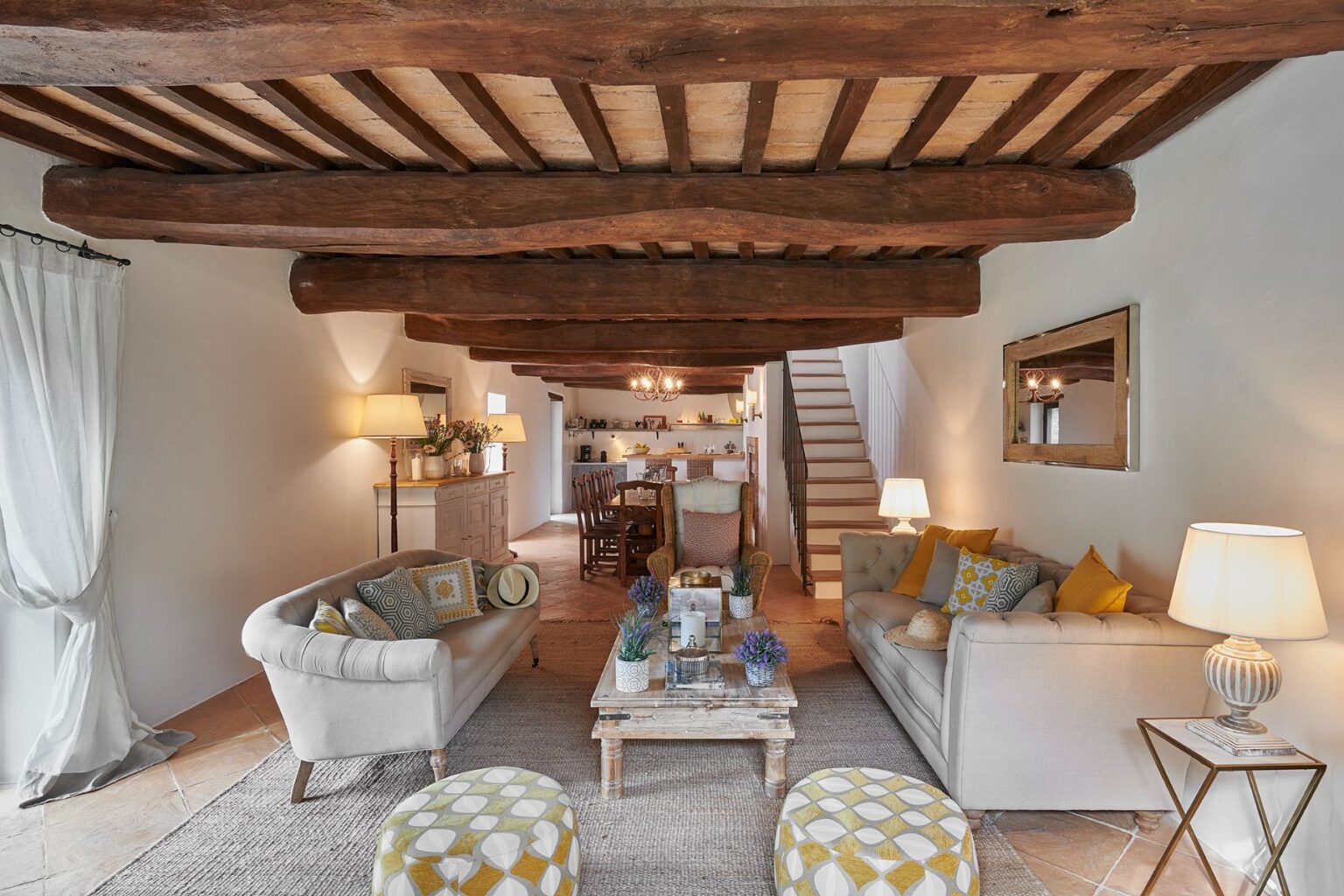 Cozy living room interior featuring wooden beams, two stylish sofas with decorative pillows, and a rustic coffee table adorned with plants. Soft lighting from floor lamps creates a warm ambiance, while a dining area is visible in the background, enhancing the inviting atmosphere. | Only on SopranoVillas