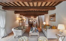 Cozy living room interior featuring wooden beams, two stylish sofas with decorative pillows, and a rustic coffee table adorned with plants. Soft lighting from floor lamps creates a warm ambiance, while a dining area is visible in the background, enhancing the inviting atmosphere.
