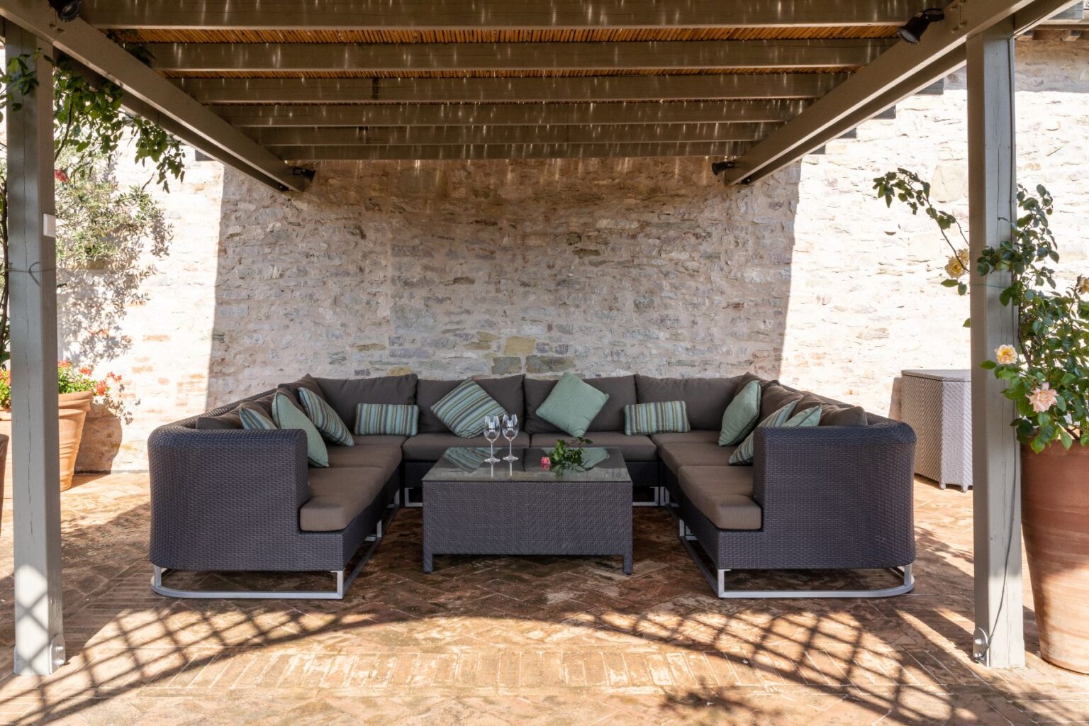 Cozy outdoor seating area with a large sectional sofa, decorative cushions, and a glass-top coffee table, surrounded by stone walls and greenery, ideal for relaxation and entertaining. | Only on SopranoVillas