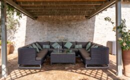 Cozy outdoor seating area with a large sectional sofa, decorative cushions, and a glass-top coffee table, surrounded by stone walls and greenery, ideal for relaxation and entertaining.