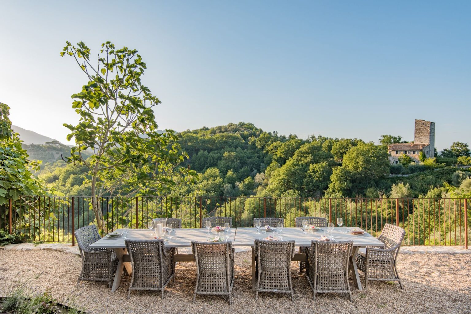Outdoor dining area with a large wooden table surrounded by wicker chairs, set against a backdrop of lush green hills and a historic stone tower. The scene captures a serene, inviting atmosphere perfect for gatherings in nature. | Only on SopranoVillas