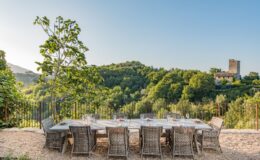 Outdoor dining area with a large wooden table surrounded by wicker chairs, set against a backdrop of lush green hills and a historic stone tower. The scene captures a serene, inviting atmosphere perfect for gatherings in nature.