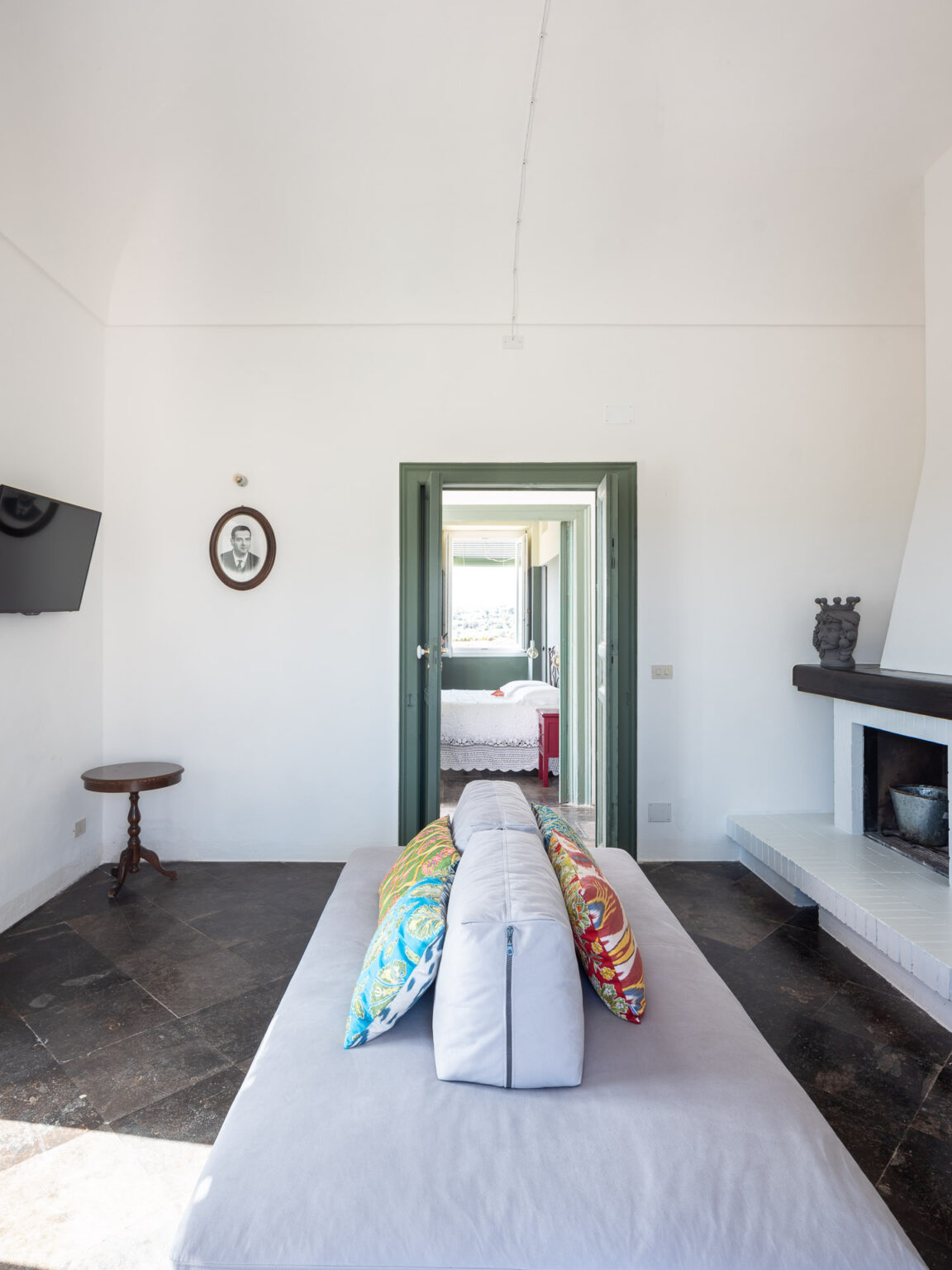 Interior view of a modern, minimalist living space featuring a comfortable sofa with colorful pillows, a round wooden table, and a television mounted on the wall. A doorway leads to a bright bedroom with a bed dressed in white linens. The room has a clean, airy aesthetic with white walls and natural light streaming in. | Only on SopranoVillas