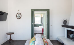 Interior view of a modern, minimalist living space featuring a comfortable sofa with colorful pillows, a round wooden table, and a television mounted on the wall. A doorway leads to a bright bedroom with a bed dressed in white linens. The room has a clean, airy aesthetic with white walls and natural light streaming in.