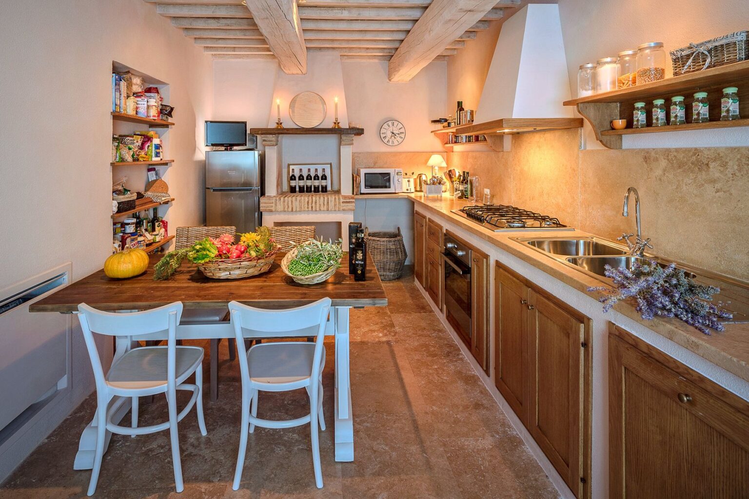 Cozy kitchen interior featuring a wooden dining table with fresh vegetables, white chairs, and open shelving stocked with pantry items. The kitchen is equipped with modern appliances, including a refrigerator and a gas stove, complemented by warm lighting and rustic decor elements. | Only on SopranoVillas