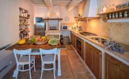 Cozy kitchen interior featuring a wooden dining table with fresh vegetables, white chairs, and open shelving stocked with pantry items. The kitchen is equipped with modern appliances, including a refrigerator and a gas stove, complemented by warm lighting and rustic decor elements.