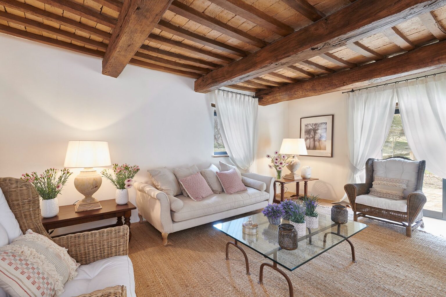 Cozy living room featuring rustic wooden beams, a light-colored sofa with decorative pillows, a wicker chair, and a glass coffee table adorned with flowers and decorative items. Natural light filters through sheer curtains, creating a warm and inviting atmosphere. | Only on SopranoVillas