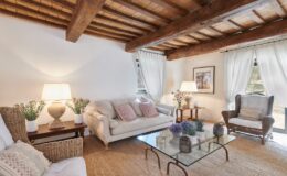 Cozy living room featuring rustic wooden beams, a light-colored sofa with decorative pillows, a wicker chair, and a glass coffee table adorned with flowers and decorative items. Natural light filters through sheer curtains, creating a warm and inviting atmosphere.